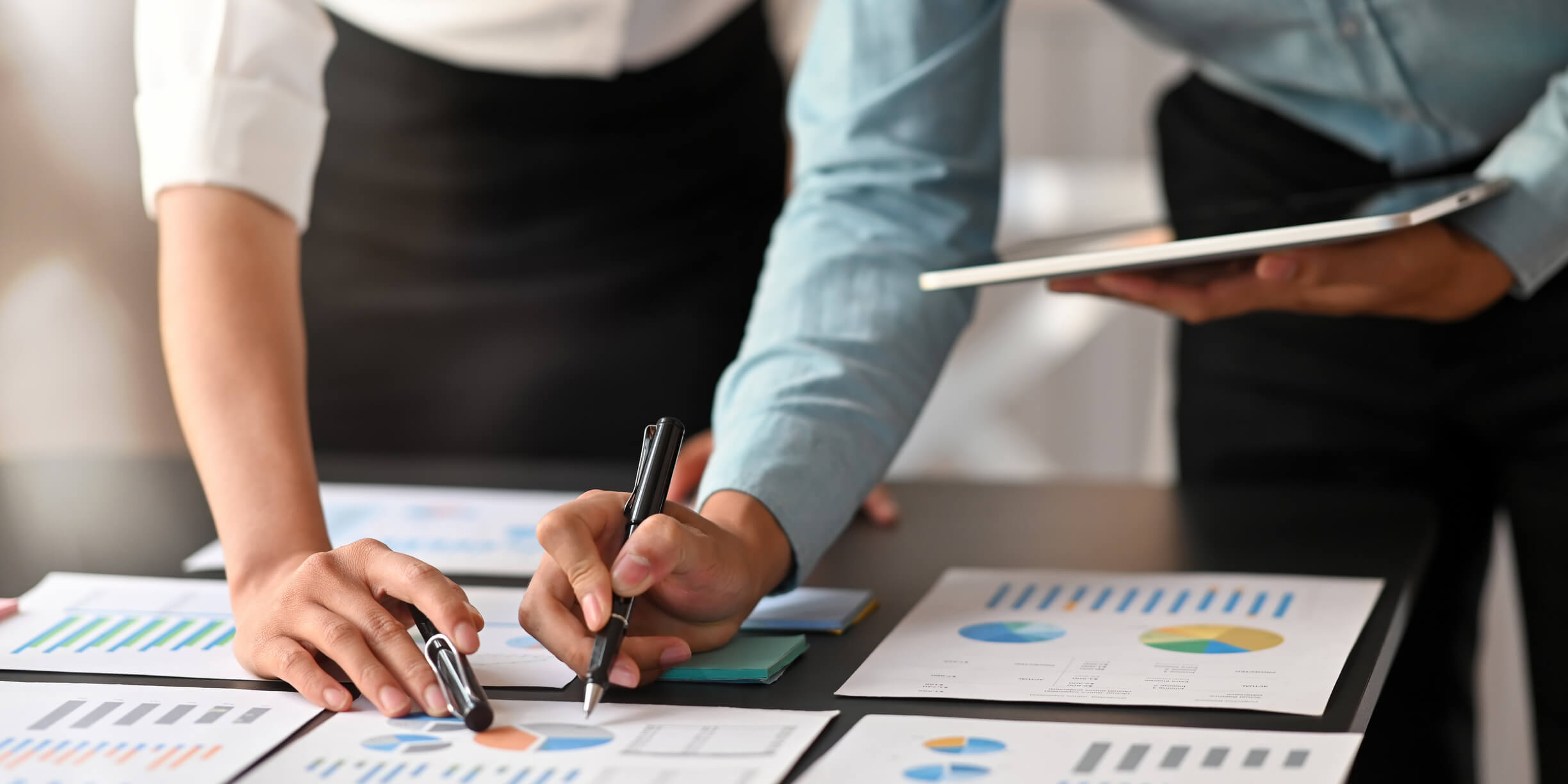 Hands of two members of a deal team taking notes on documents with charts and graphs to perform an analysis